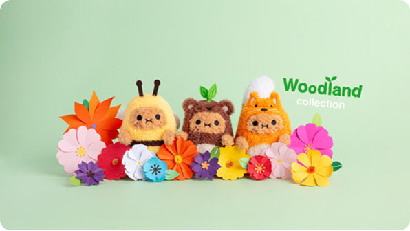 Three plush woodland animal toys, including a yellow bee, a brown bear, and an orange fox, arranged with colorful paper flowers on a light green background. The text 'Woodland collection' is visible on the right.