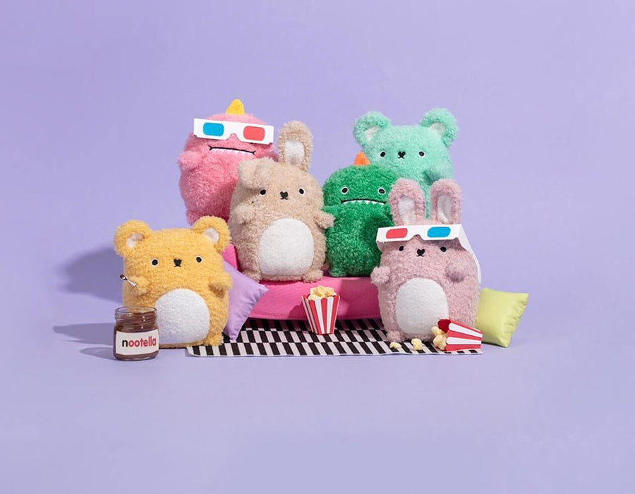 A group of cute plush animal toys arranged on a pink couch with popcorn and a jar of Nootropil, set against a solid lavender background.