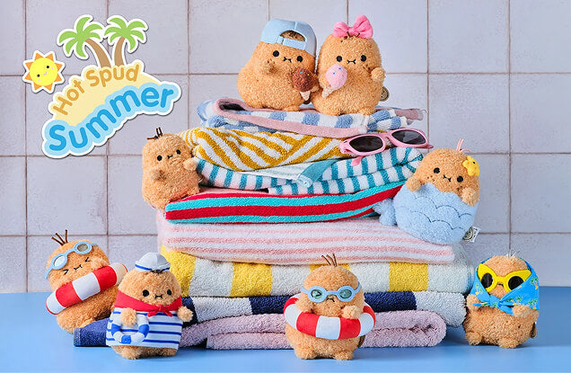 A stack of plush Hot Spud Summer-themed towels and matching plush toys arranged on a blue surface against a light tiled wall. The set includes striped towels in pink, yellow, blue, and white, along with small brown plush figures wearing swim hats, sunglasses, life preservers, and tropical outfits.